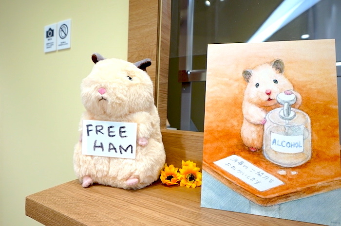 右：エントランスにて見つけた、手指の消毒をお願いする助六。左：「FREE HUG」ならぬ「FREE HUM」の札を掲げたぬいぐるみは、ショップで購入することができる（税込3,300円）