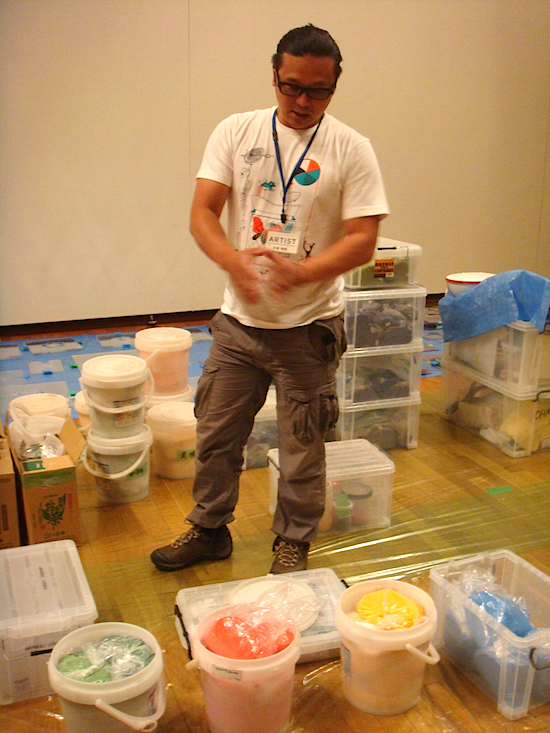 顔料について説明する大巻伸嗣。これまでの作品で使用したストックに、今回のために用意した300kgを加え、総重量1トンもの顔料を用意。過去のストックの中にはもう入手不可能な色もあるそうで、色数は100種類以上にも及ぶ