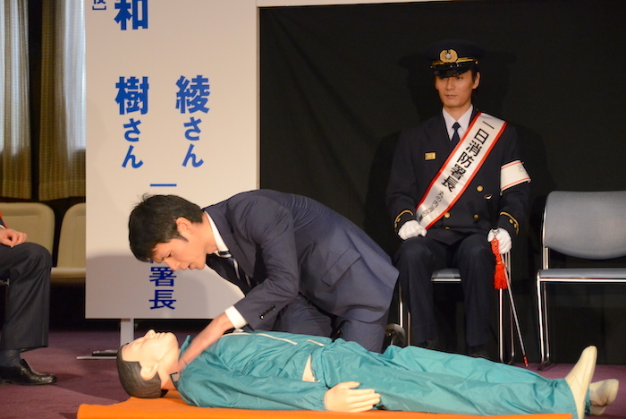 AEDの使い方のデモンストレーションの様子を眺める加藤和樹(右奥)