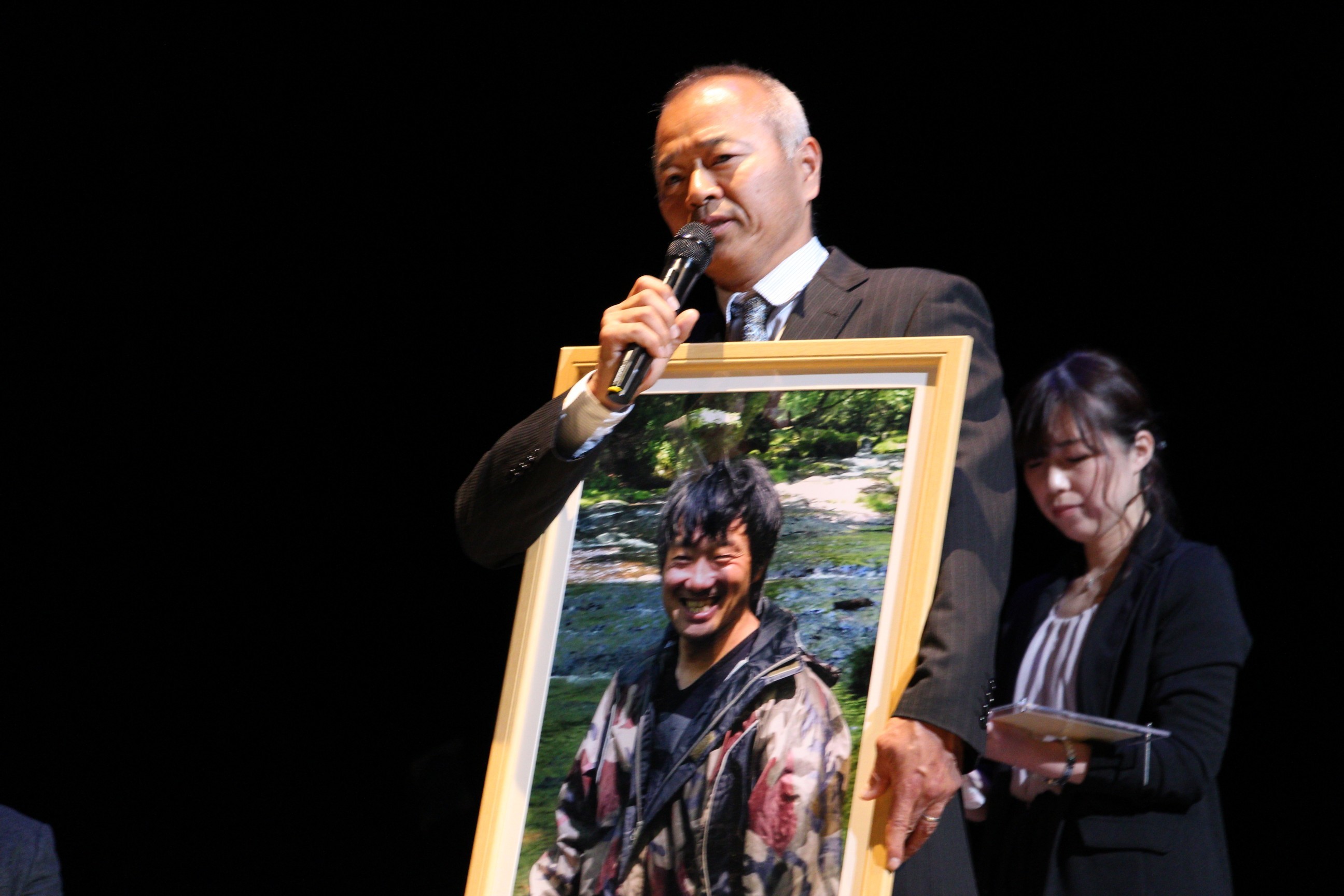 左から、舟山弘一さんの写真を掲げる辻井啓伺氏、舟山さんの妻・めぐみさん